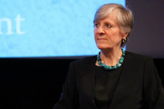 A woman wearing a turquoise necklace sits on stage during a panel discussion with a presentation screen visible in the background.