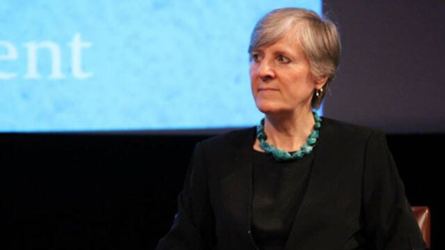 A woman wearing a turquoise necklace sits on stage during a panel discussion with a presentation screen visible in the background.