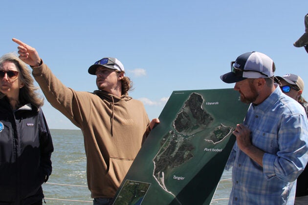 A group of people examine a map showing the erosion of Tangier Island while standing along the shoreline.