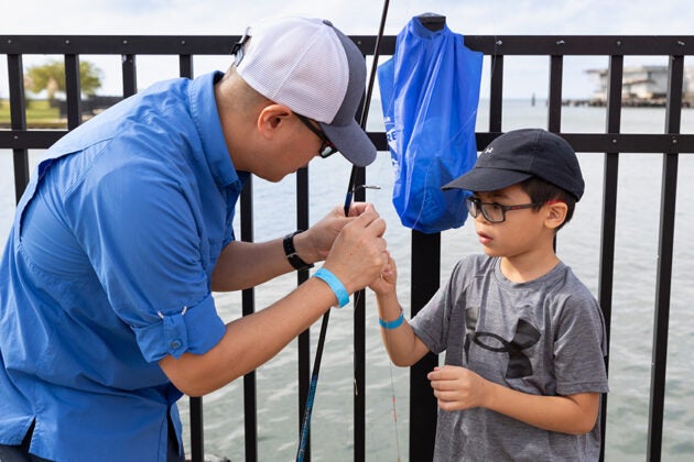 An adult helps a young boy prepare fishing tackle at a waterfront railing.