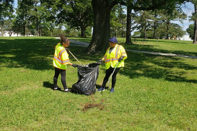 Two volunteers in yellow safety vests collect litter into a trash bag in a park.