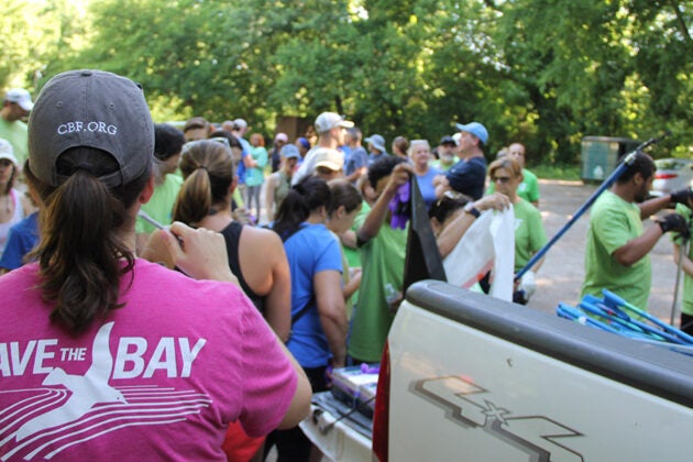 A volunteer wearing a Save the Bay shirt and CBF cap stands near a pickup truck as a crowd gathers for a cleanup event.