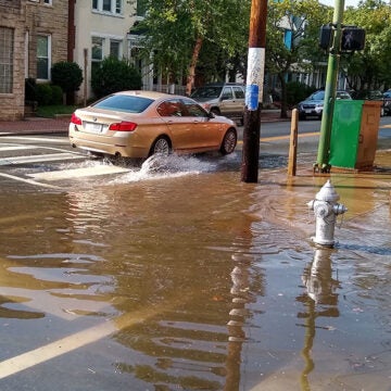 A car drives through floodwater on a city street, with a fire hydrant partially submerged at the intersection.