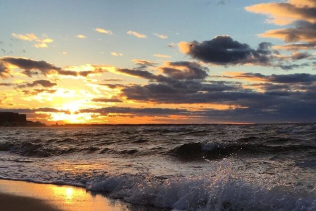 Dramatic sunset over Virginia Beach with golden clouds, rolling waves, and silhouetted buildings along the shore.