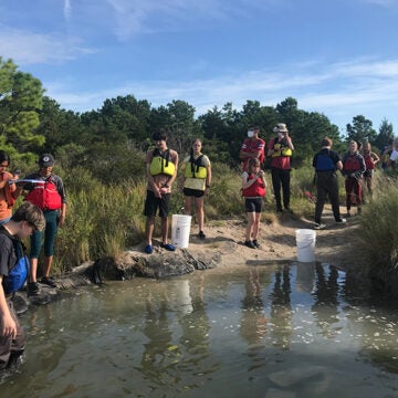 A dozen students stand with their teacher on the shore of a creek.
