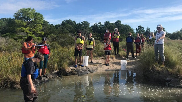 A dozen students stand with their teacher on the shore of a creek.