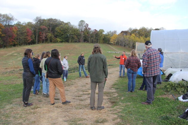 Local farmers, elected officials, nonprofit partners, and students stand gathered in a group next to a greenhouse at the Regenerative Farm Tour in Frederick
