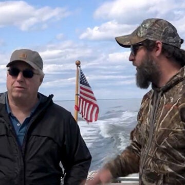 Two men, one in a blue jacket and one in a camouflage jacket talk on the stern of a boat, with an American flag and the boat's wake seen behind them.