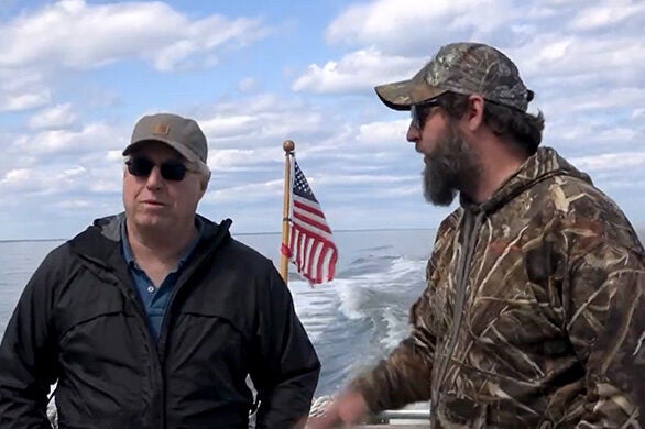 Two men, one in a blue jacket and one in a camouflage jacket talk on the stern of a boat, with an American flag and the boat's wake seen behind them.