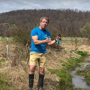 A man in a blue CBF shirt stands next to a small stream running through a field.