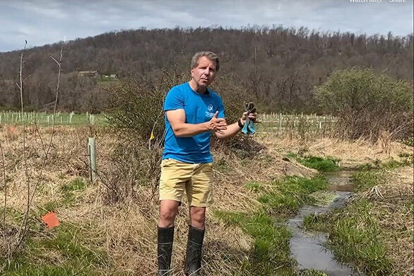 A man in a blue CBF shirt stands next to a small stream running through a field.