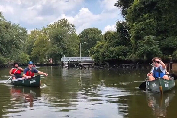 Students in blue and red personal flotation devices paddle a stream in green canoes with the names Raccoon and Red Fox.