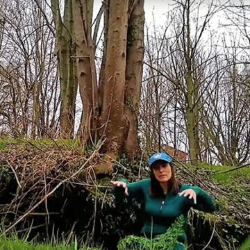 A woman in a green jacket and CBF cap sits on the ground in front of trees.