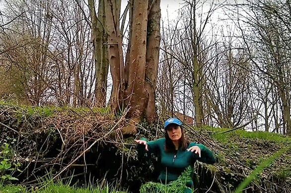 A woman in a green jacket and CBF cap sits on the ground in front of trees.