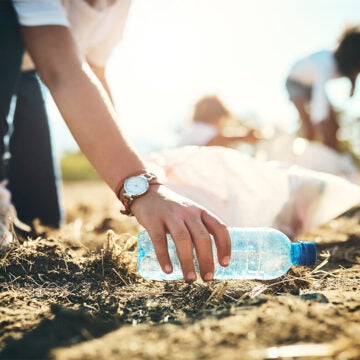 A hand reaches down and picks a plastic bottle up off the sand. Text reads Clean the Bay Day.