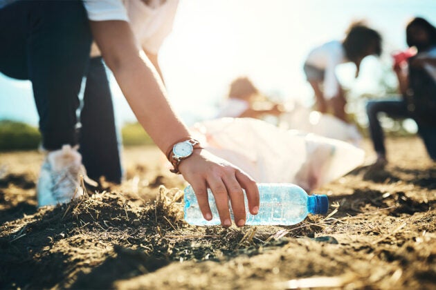 A hand reaches down and picks a plastic bottle up off the sand. Text reads Clean the Bay Day.