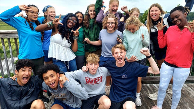 A joyful group of students cheer and strike playful poses together on a windswept boardwalk, celebrating the moment.