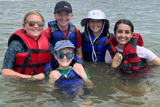 Five young girls in life jackets, grinning ear to ear as they stand in the Bay.