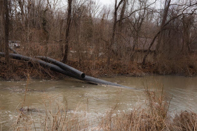 Large pipes drape through woods into a brown river.