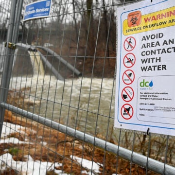 Sewage filled water spills from pipes into a river behind a fence bearing a warning sign.