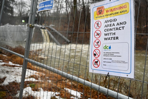 Sewage filled water spills from pipes into a river behind a fence bearing a warning sign.