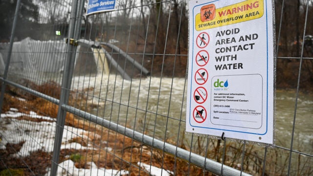 Sewage filled water spills from pipes into a river behind a fence bearing a warning sign.
