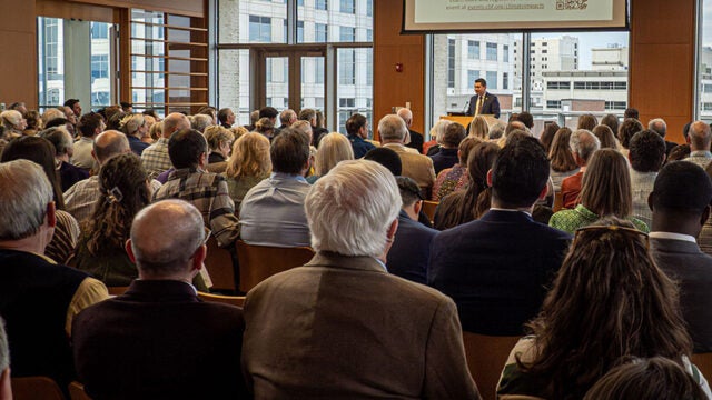 A group of seated people watch a man at a podium, a PowerPoint presentation displayed on a screen above him.