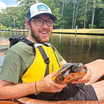 Connor Trexel wears a yellow vest and holds a turtle while sitting in an orange kayak.