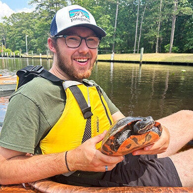 Connor Trexel wears a yellow vest and holds a turtle while sitting in an orange kayak.