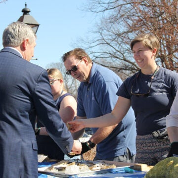 A smiling woman hands a man a newly shucked oyster while others stand next to her at a table shucking more oysters.