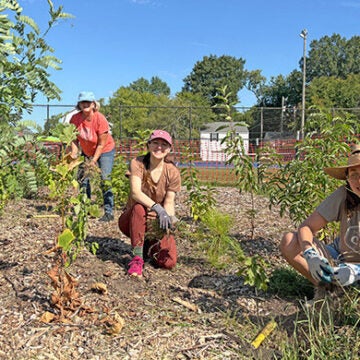 Three women working amid newly planted trees and shrubs smile at the camera.