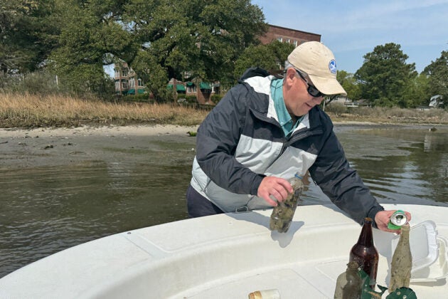 A man standing in the water places old bottles, food cartons, and other trash on the deck of a boat.