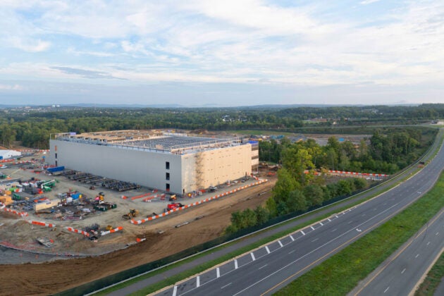 A large commercial building is under construction beside a busy highway, surrounded by equipment, vehicles, and open landscape.