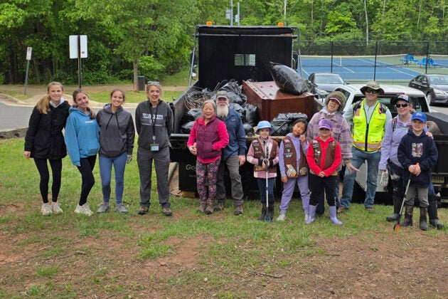 A line of smiling people stand in front of a dump truck full of trash bags and a dresser.