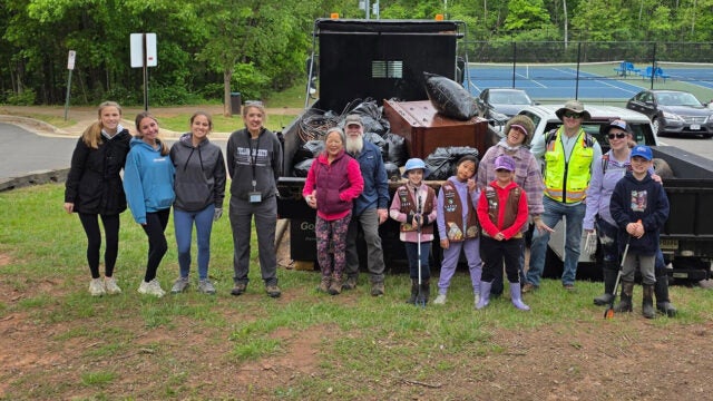 A line of smiling people stand in front of a dump truck full of trash bags and a dresser.