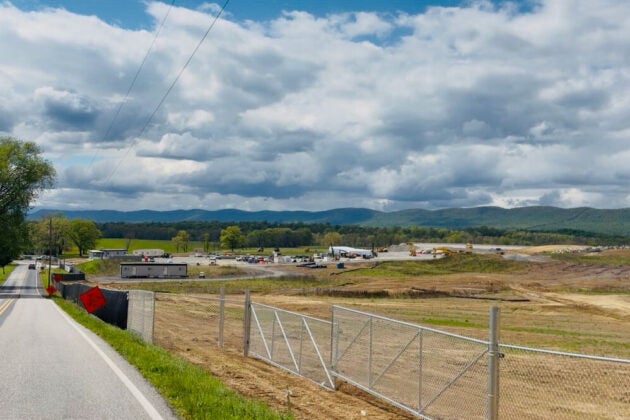 Photo of a large commercial building under construction beside a residential road, surrounded by equipment, vehicles, and open landscape.