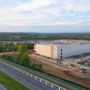 A large industrial building under construction sits beside a multilane highway, surrounded by construction equipment, orange barriers, and a wide, partly cloudy sky.