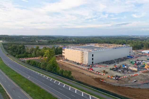 A large industrial building under construction sits beside a multilane highway, surrounded by construction equipment, orange barriers, and a wide, partly cloudy sky.