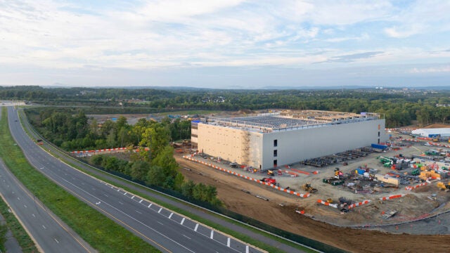 A large industrial building under construction sits beside a multilane highway, surrounded by construction equipment, orange barriers, and a wide, partly cloudy sky.