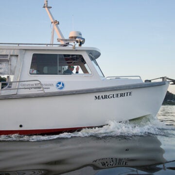 A white boat named “MARGUERITE” with a Chesapeake Bay Foundation logo cruises on calm water while a person wearing sunglasses waves from the cabin.