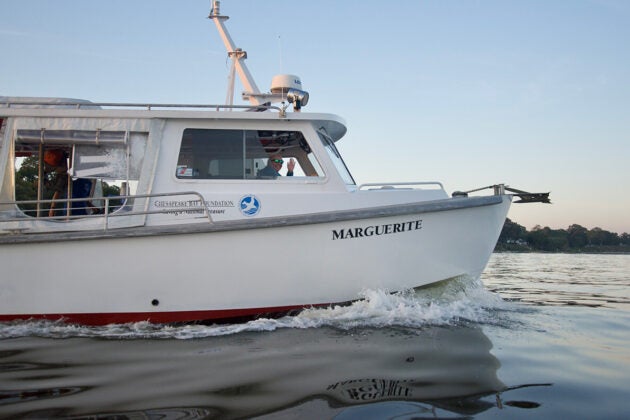 A white boat named “MARGUERITE” with a Chesapeake Bay Foundation logo cruises on calm water while a person wearing sunglasses waves from the cabin.