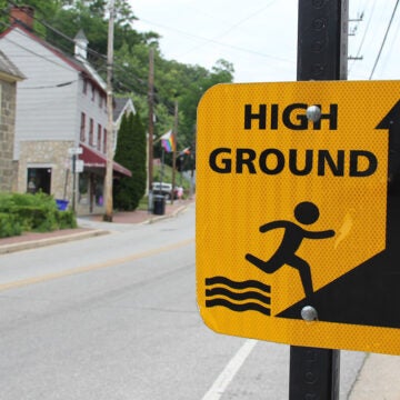 A close-up of a yellow “HIGH GROUND” warning sign mounted on a pole with a small-town street, buildings, and a rainbow flag visible in the background.