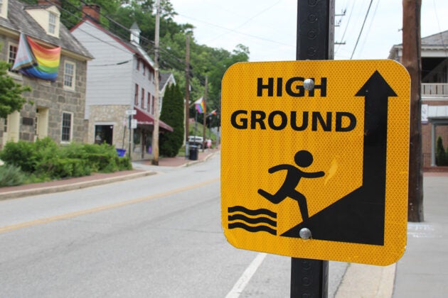 A close-up of a yellow “HIGH GROUND” warning sign mounted on a pole with a small-town street, buildings, and a rainbow flag visible in the background.