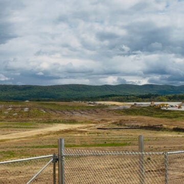 A wide-open construction site behind a chain-link fence stretches toward green rolling hills under a dramatic, cloud-filled sky.