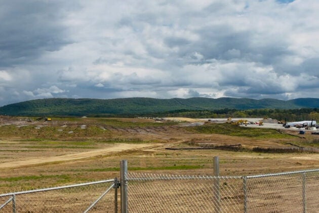 A wide-open construction site behind a chain-link fence stretches toward green rolling hills under a dramatic, cloud-filled sky.
