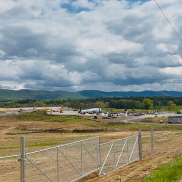 A wide-open construction site behind a chain-link fence stretches toward green rolling hills under a dramatic, cloud-filled sky.