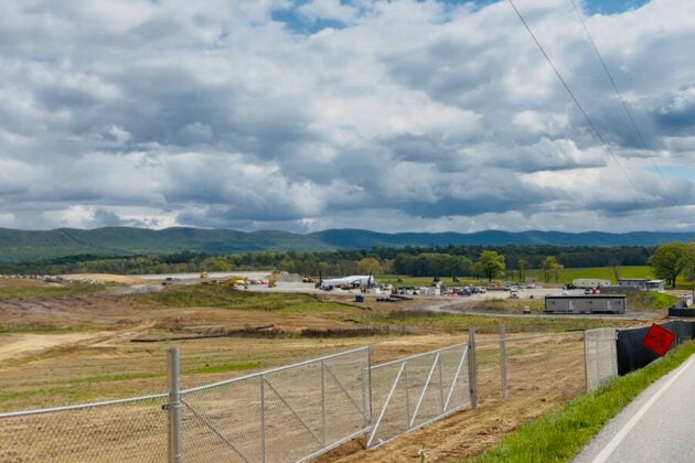 A wide-open construction site behind a chain-link fence stretches toward green rolling hills under a dramatic, cloud-filled sky.