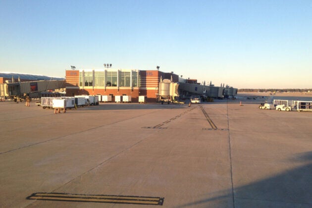 Several building and airport vehicles are visible across a runway with a light blue sky in the background.