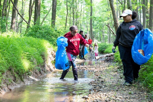 A girl in a red Harvard sweatshirt and high boots walks across a woodland creek carrying a blue trash bag while another woman with a trashbag stands on the creekside waiting for her.