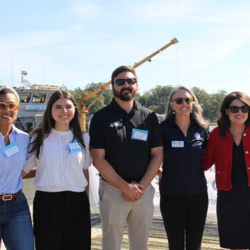 Five people stand side by side outdoors on a dock near water, smiling at the camera with a boat and trees in the background.